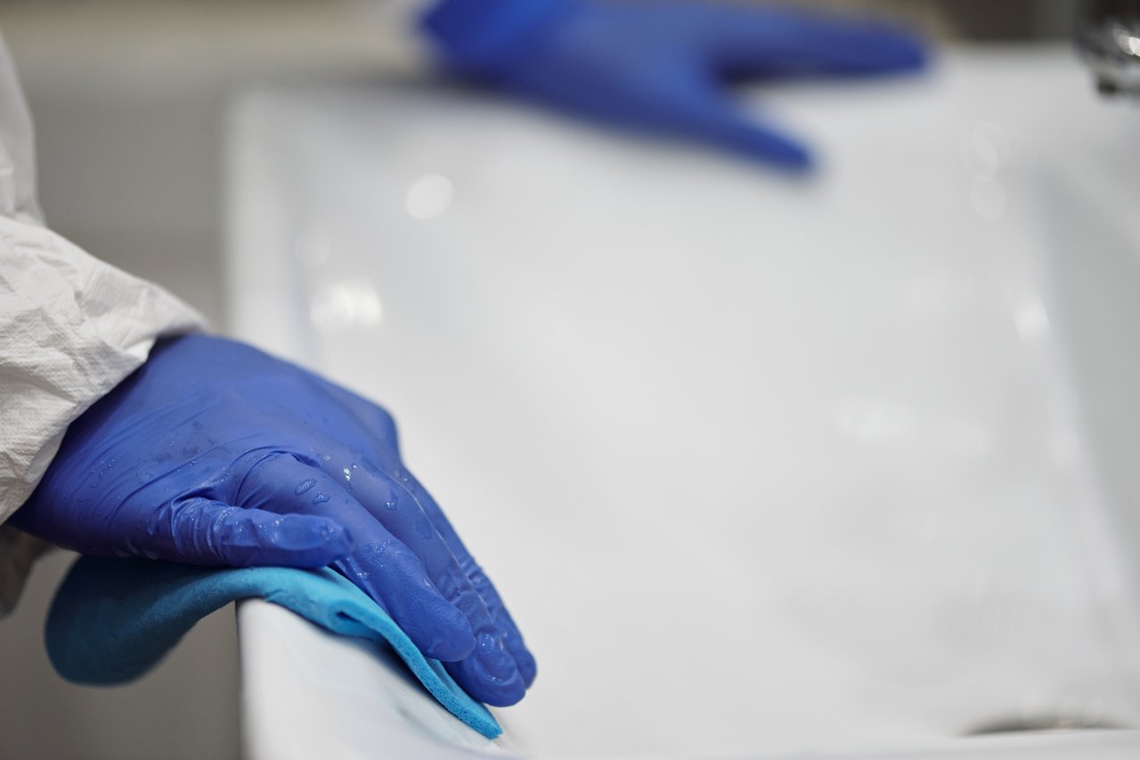 hero-img Close-up of a person sanitizing a bathroom sink with gloves and cloth, focusing on hygiene and cleanliness.